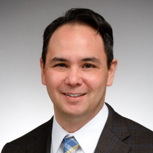 Professional headshot of a smiling man with short dark hair wearing a suit jacket, white shirt, and patterned tie against a neutral background.