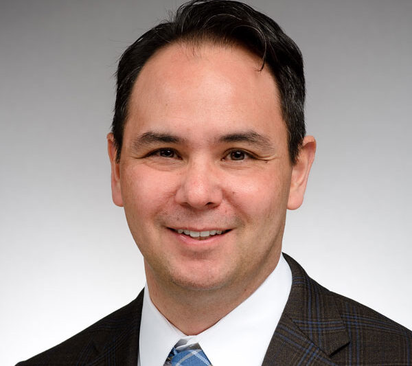 Professional headshot of a smiling man with short dark hair wearing a suit jacket, white shirt, and patterned tie against a neutral background.