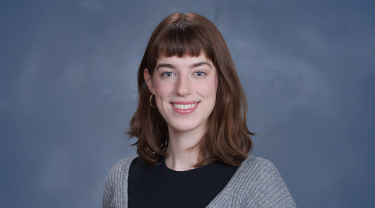 A young woman with auburn hair wearing a black shirt and gray sweater stands against a blue background and smiles