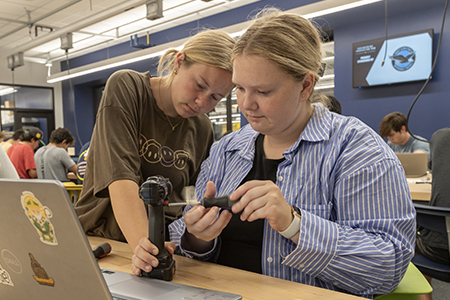 Two students work together to take apart a power drill, referencing their laptop as they examine its components.