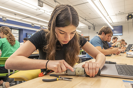A student focuses on loosening a screw from a disassembled power drill, with tools and a laptop nearby.