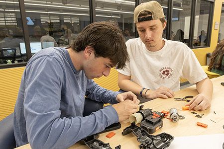 Two students carefully dismantle a power drill, organizing the pieces on the table.