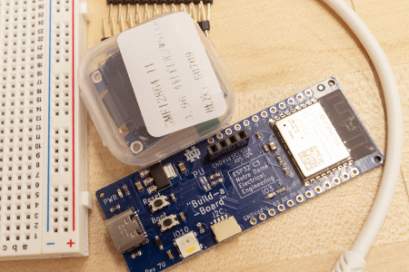 A blue circuit board labeled "Build-a-Board" lies on a work surface next to a breadboard and electronic components.