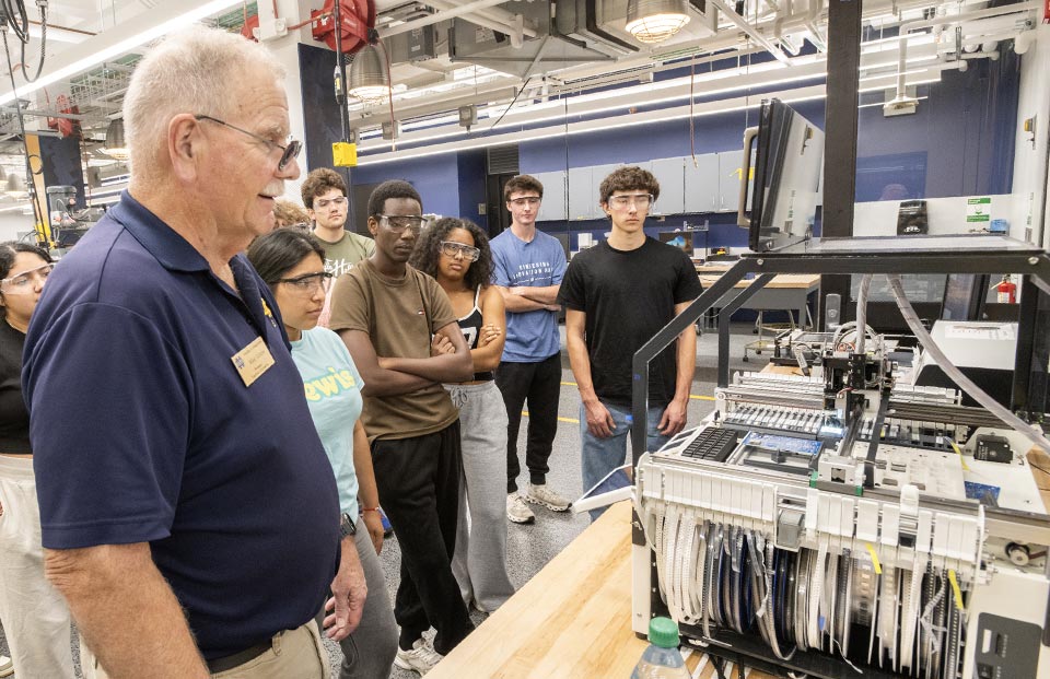 Instructor Mike Schafer explains the circuit board assembly process to a group of students gathered around a machine in the Engineering Innovation Hub.