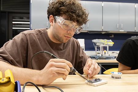 A student wearing safety goggles carefully solders parts onto a circuit board while seated at a workbench.