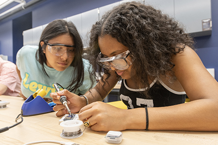 Two students wearing safety goggles work together with a soldering iron to attach components to a small board.