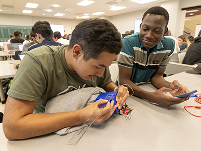 A student focuses on adjusting wiring inside a blue sensor housing while a teammate observes and holds a phone.
