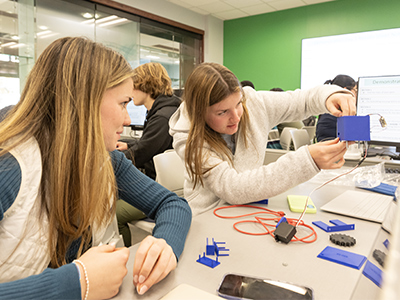 Two students examine and discuss the wiring of a blue sensor housing at their lab bench, with design components laid out nearby.
