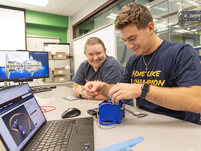 Two students sit at a table smiling while working together on a small blue device next to a laptop displaying CAD software.