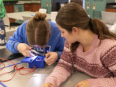 Two students collaborate at a lab table, inspecting a wired sensor prototype with tools and components spread out.