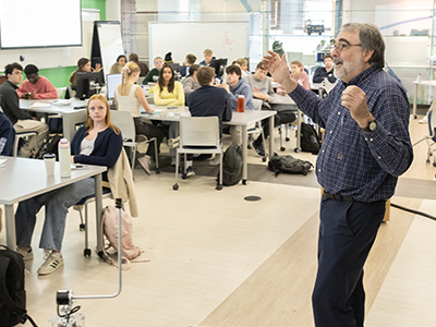 EE department chair Greg Snider gestures while speaking to a classroom of first-year students seated at round tables.