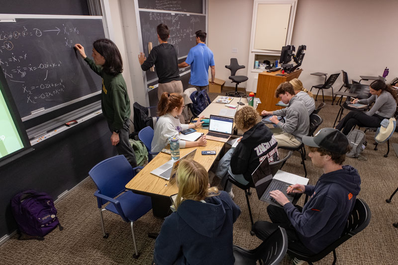 A young man writes on the blackboard while a group of students study at tables in front of him.