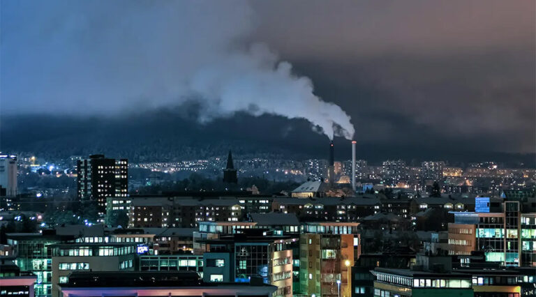 Nighttime city view with many lit buildings. Two industrial chimneys, one black and one white, glow red at their tops while releasing thick white smoke into the dark, cloudy sky.