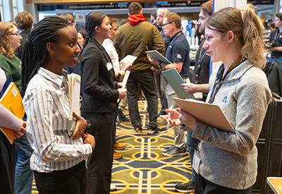 Student speaking with a recruiter while holding a folder in a crowded career fair hall.