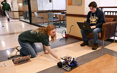 Student kneeling on the floor adjusting a small robotic device while another monitors it from a laptop nearby.