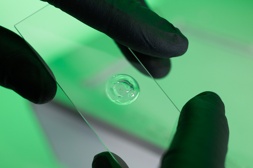 A small transparent droplet of biomaterial rested on a glass slide, held steady by gloved hands under green-tinted lighting.