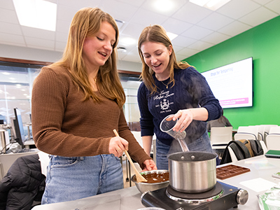 Two students smile and work together, stirring and pouring chocolate into a pot as part of a hands-on lab activity.