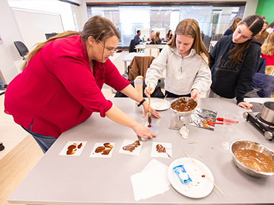An instructor guides a small group of students as they spread and shape chocolate samples on a table, observing technique and results.