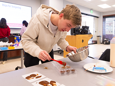 A student leans over a table, spooning melted chocolate from a metal bowl onto small samples with focused attention.