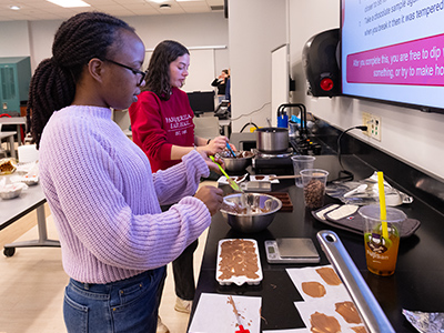 Students work at a lab bench preparing chocolate mixtures, measuring and spreading samples while following a recipe in a classroom setting.