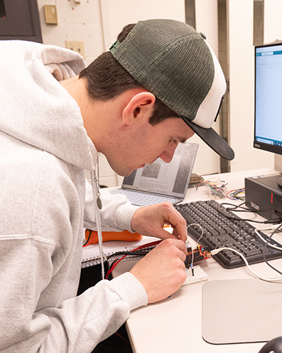 A student works at a desk assembling a small electronic circuit, carefully placing wires into a breadboard beside a computer.