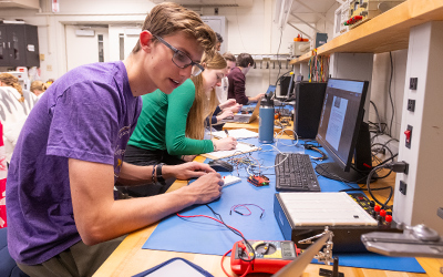 A student in a lab workspace connects wires and components while referencing a computer, working alongside classmates at a bench.