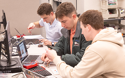 Three students sit together at a workstation, collaborating on an electronics project using laptops and circuit components.