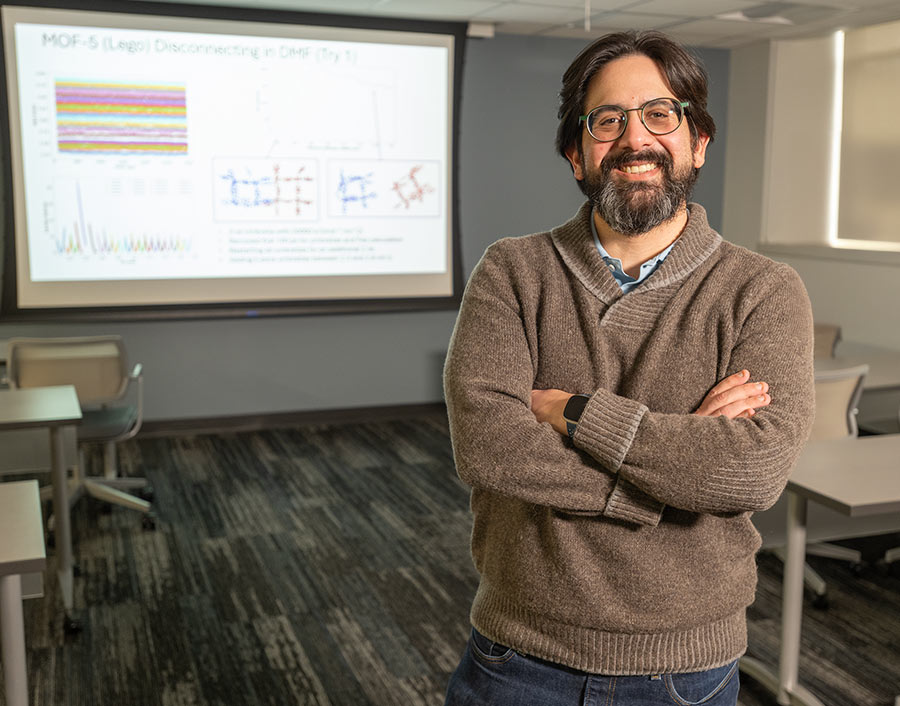 A faculty member stands in a classroom with arms crossed, smiling at the camera, with a presentation slide of colorful scientific data displayed on a screen behind him.