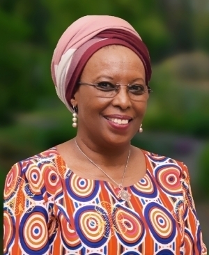 Smiling woman in a pink and burgundy headwrap, glasses, and a vibrant orange, blue, and red patterned top.