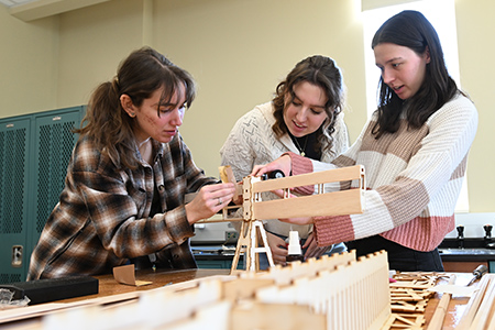Three students collaborate to assemble a wooden aircraft frame at a lab table.