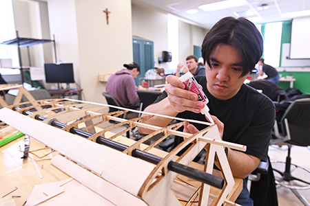Student applies glue to a detailed wooden aircraft wing structure in a workspace.
