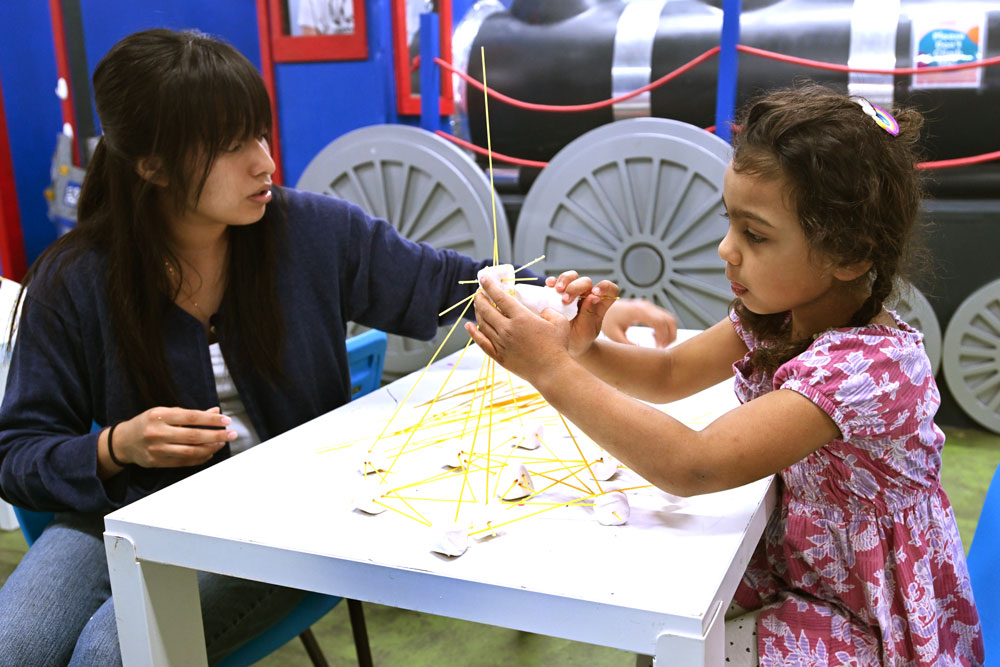 Student assists a child building a small marshmallow-and-spaghetti noodle structure at a table.