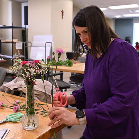 Woman trims flower stems with scissors while arranging a bouquet at a table.