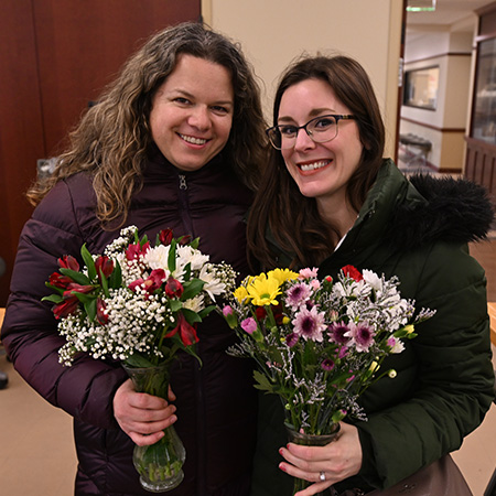 Two women smile while holding colorful flower bouquets in glass vases.