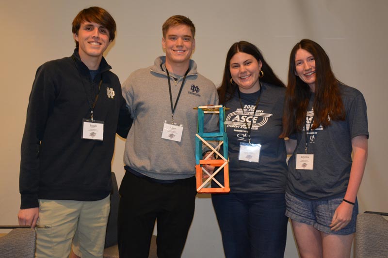 Four Notre Dame students stand together indoors, smiling and holding a small model structure built for the Mystery Design Competition focused on earthquake engineering.