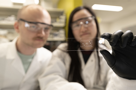 Two researchers in lab coats examine a small transparent chip while wearing safety goggles.