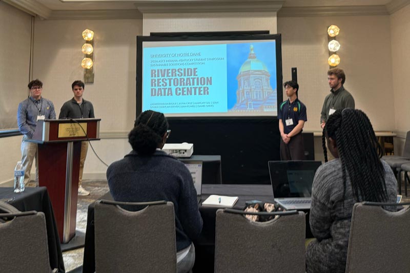 Four students present a project at the front of a room while judges sit facing them, with a slide titled “Riverside Restoration Data Center,” representing Notre Dame’s second-place finish in the Sustainable Solutions Competition.