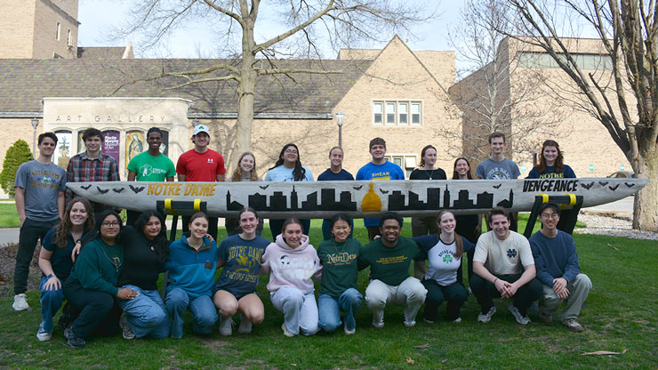 A large group of Notre Dame students pose outdoors in front of a building, holding a long concrete canoe decorated with a city skyline design, marking their second-place finish in the Concrete Canoe Competition.