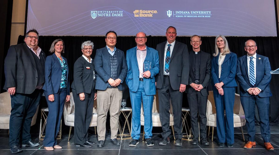 Nine people, including a man holding a crystal award and a man in a clerical collar, smile on a stage.