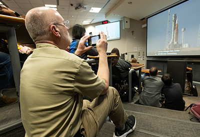 Professor Clive Neal sits on the steps of a lecture hall photographing the Artemis II launch broadcast as students watch the livestream projected at the front of the room.