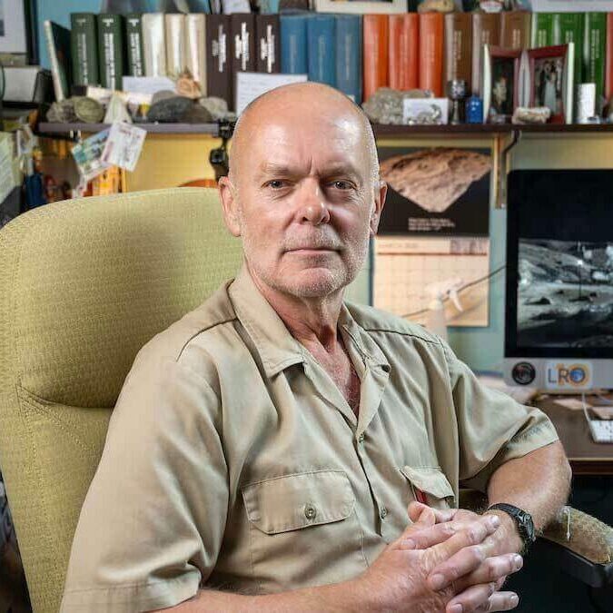 Clive Neal seated in his office at Notre Dame, with a lunar surface image on the computer monitor behind him and shelves of research materials above.