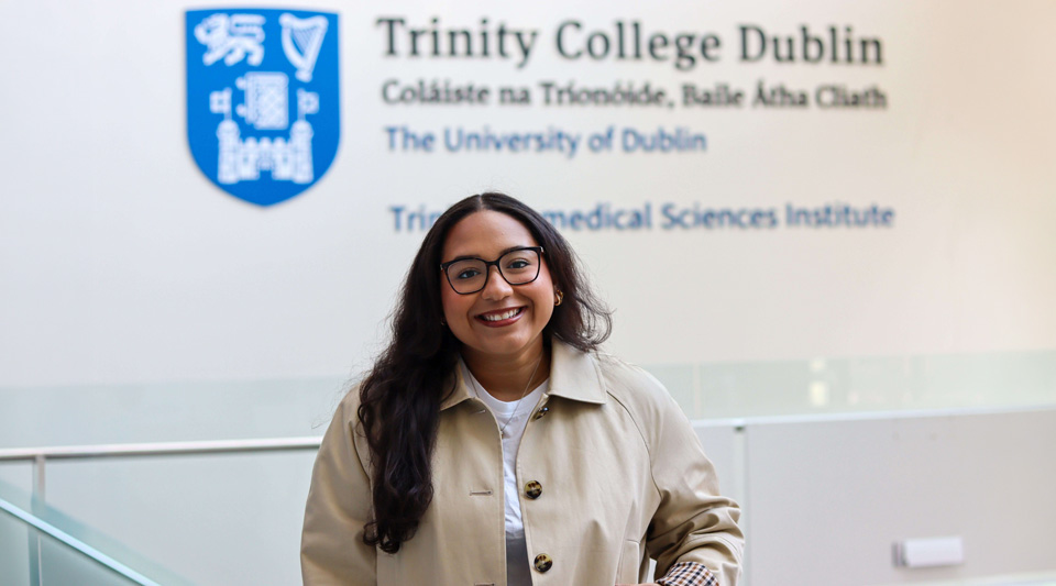 Smiling woman with dark hair and glasses in a tan coat, standing before the Trinity College Dublin wall logo and text.