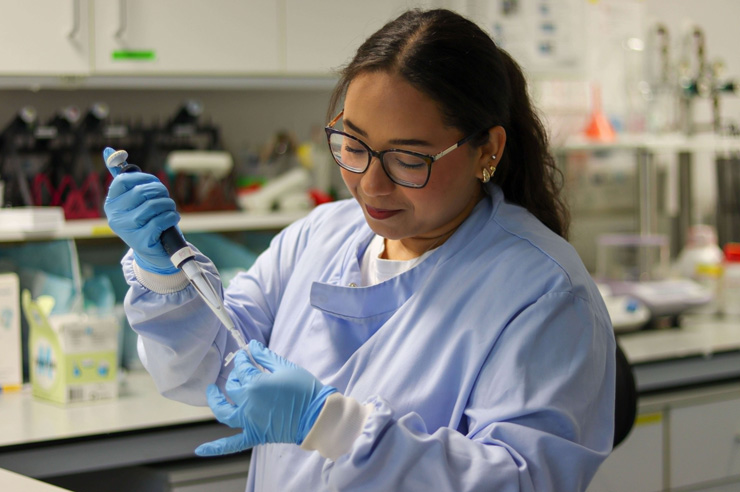 Woman wearing glasses, blue lab coat, and gloves intently pipetting samples in a science lab.