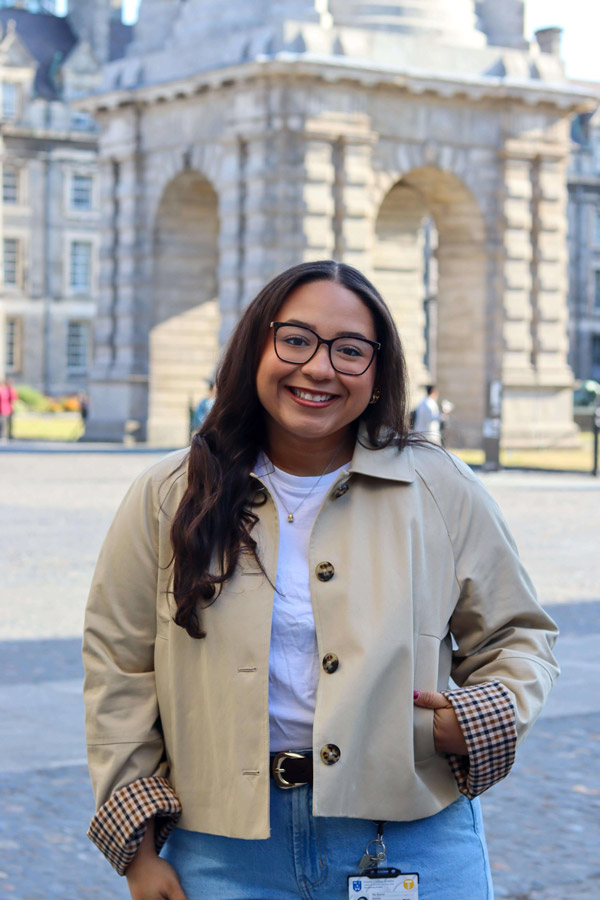 Smiling woman in glasses, beige jacket with plaid cuffs, stands before historic stone arches at Trinity College Dublin.