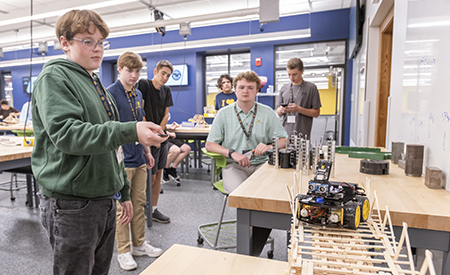 A high school student operates a robotic car as other students watch it cross a wooden bridge.