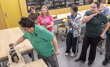 An instructor tests a student-built wooden bridge while high school students look on.