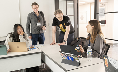 Students and faculty gather around a table with laptops during a collaborative research session.