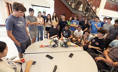 Students gather around a table to watch robots during a hands-on engineering activity.