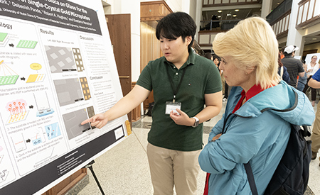 A student presents research findings to an attendee beside a research poster.