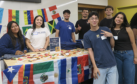 Students from an engineering organization pose at an information table featuring international flags.